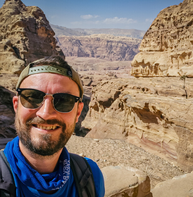 A person wearing sunglasses and a cap smiles, taking a selfie. The backdrop features towering, rugged sandstone formations under a clear blue sky.