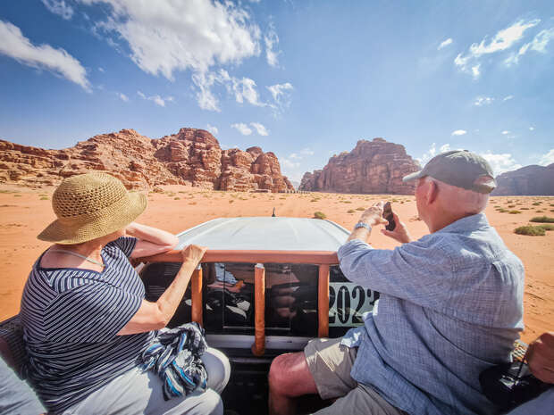 Two people sit on a vehicle photographing rugged sandstone formations in a sunlit desert landscape. Text on the vehicle reads "2023." Sparse vegetation is visible under a bright blue sky.