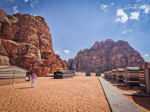 A person walks through a desert camp with striped tents, surrounded by towering red rock formations under a clear blue sky.