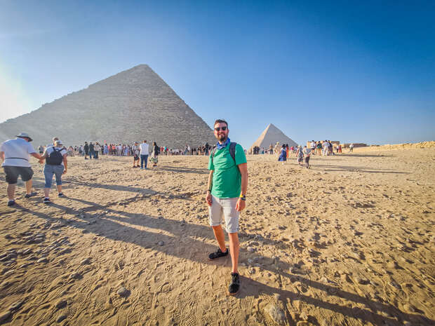 A man stands smiling in front of the pyramids. He's wearing a green shirt and shorts. Tourists and the desert environment surround him under a clear blue sky.