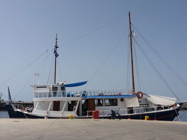 A white and blue boat docked at a harbor, featuring two tall masts and a blue flag. The sunlit deck has railings and a lifebuoy. The sky is clear.