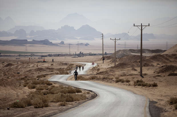 A cyclist rides on a winding, paved road flanked by power lines in a vast, barren desert landscape, with distant mountains blurred by haze.