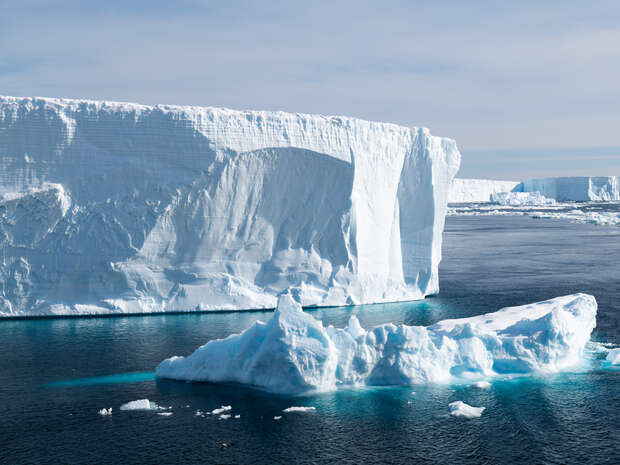 A large iceberg floats serenely on dark blue water, set against a massive ice shelf under a cloudy sky, indicating an Arctic or Antarctic setting.