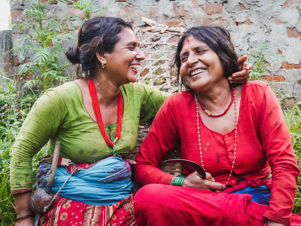 Two women, one in green and the other in red, sit laughing together. They are amidst greenery and a textured brick wall, suggesting a casual, outdoor setting.