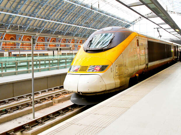 A yellow high-speed train, stationary on tracks, is positioned inside a large, glass-roofed station with arched steel beams. Passengers are not visible on the platform.