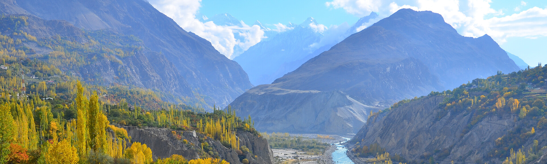 Mountains tower over a lush valley with a winding river, surrounded by vibrant autumn trees and small houses, set against a backdrop of blue sky and fluffy clouds.