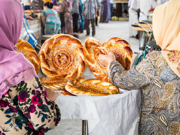 Flat, circular breads are displayed on a table with intricate designs, as two women in patterned clothing inspect them. A busy market scene unfolds in the background.