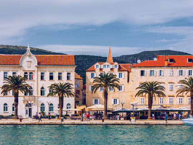 Buildings with orange-tiled roofs stand beside a waterfront, framed by palm trees. People walk along the promenade. Hills rise in the background under a partly cloudy sky.