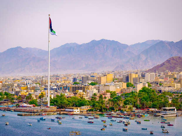 A tall flagpole stands prominently, flying a colorful flag, amidst a cityscape with numerous buildings; a bay with boats and distant mountains provide a picturesque backdrop.