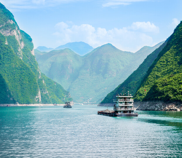 Two boats cruise through a wide river flanked by steep, lush green mountains under a bright blue sky, creating a serene and picturesque landscape.
