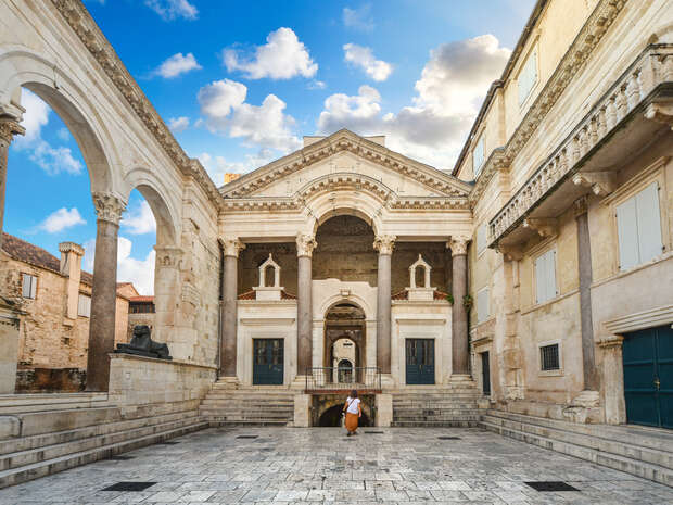 A person walks through a stone courtyard surrounded by ornate ancient architecture featuring arches and columns, under a blue sky with scattered clouds.