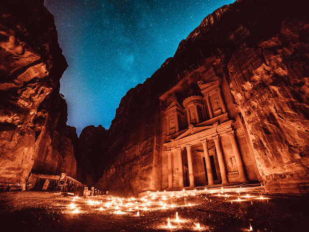 An ancient rock-carved facade, illuminated by numerous candles on the ground, stands between towering cliffs under a starry night sky.