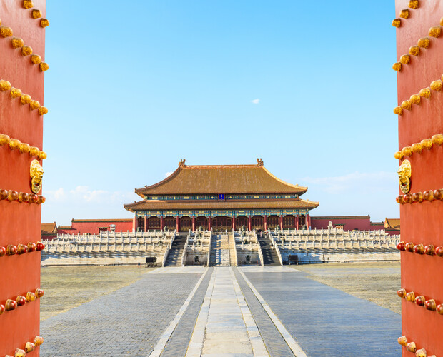 A traditional Chinese palace stands majestically within a courtyard, framed by large red doors adorned with golden studs, under a clear blue sky.