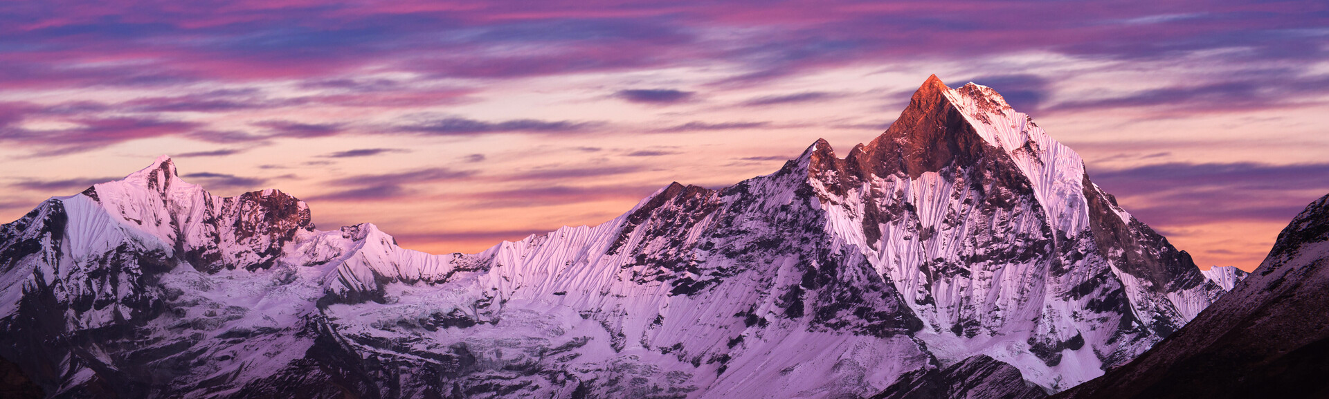 Snow-covered mountains, reflecting pink and purple hues from a sunset sky, stand majestically against a backdrop of vibrant, colorful clouds, creating a serene and picturesque landscape.