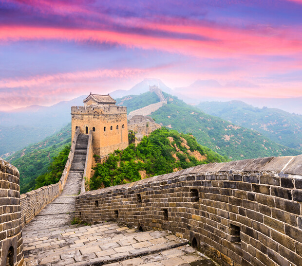 A stone walkway leads to a watchtower along a portion of the Great Wall of China, surrounded by lush green hills under a colorful, vibrant sky with pink and purple hues.