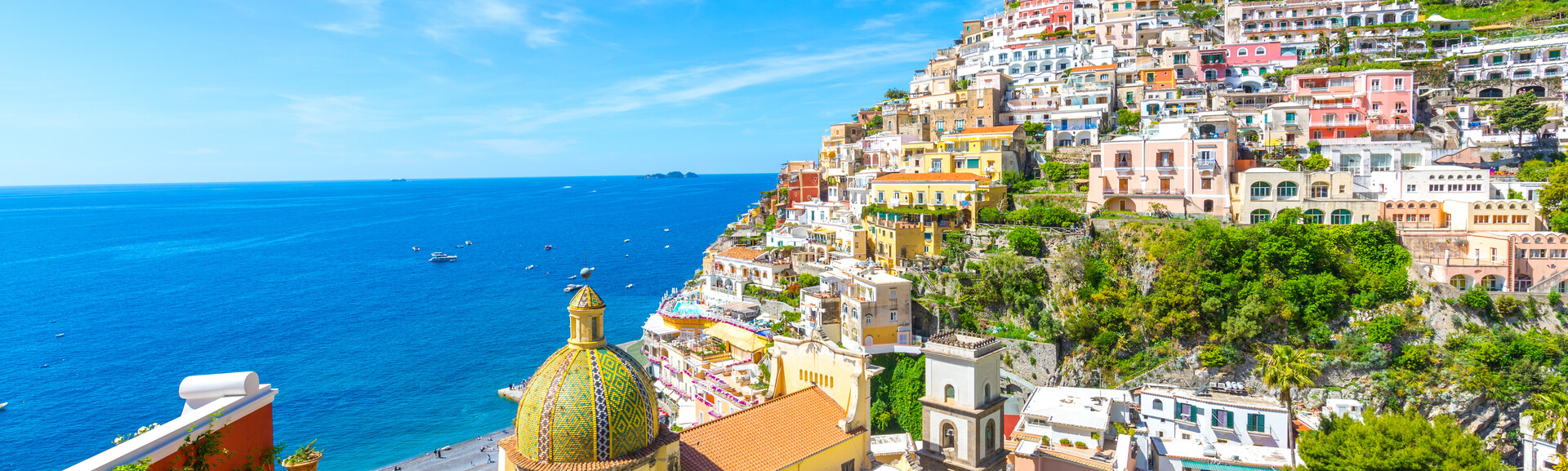 Clusters of colorful houses ascend a steep hillside, overlooking a serene blue sea with small boats scattered on the water, under a clear sky. A domed church stands prominently.