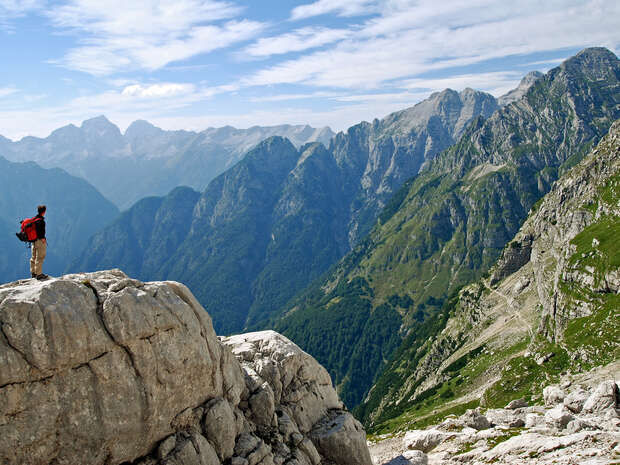 A lone hiker with a red backpack stands atop a rocky cliff, overlooking expansive, rugged mountains under a partly cloudy sky. Green vegetation covers the lower slopes beyond the rocks.