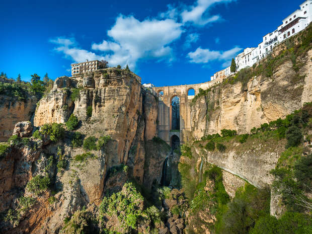 A stone bridge spans a deep gorge, connecting cliffs with houses perched above. Lush greenery accents the rugged rock faces under a bright blue, partly cloudy sky.
