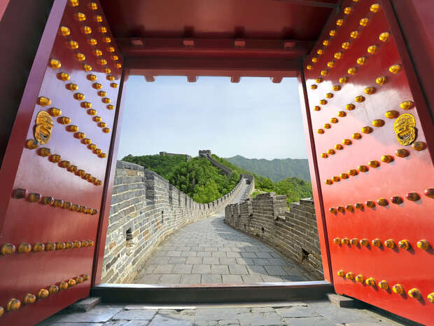 Red, heavily studded doors open onto a winding stone path of the Great Wall, set against a backdrop of green hills under a clear blue sky.