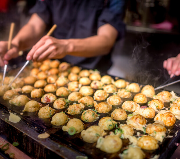 Takoyaki balls being cooked on a hot grill, with hands using skewers to turn them, surrounded by a busy kitchen setting. Steam rises, indicating the heat and freshness.