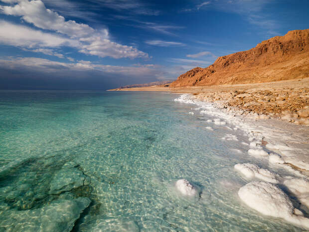 Clear turquoise water laps against a rocky shoreline with salt deposits, under a sky with scattered clouds. Rugged red-hued cliffs rise on the right, suggesting a coastal desert landscape.