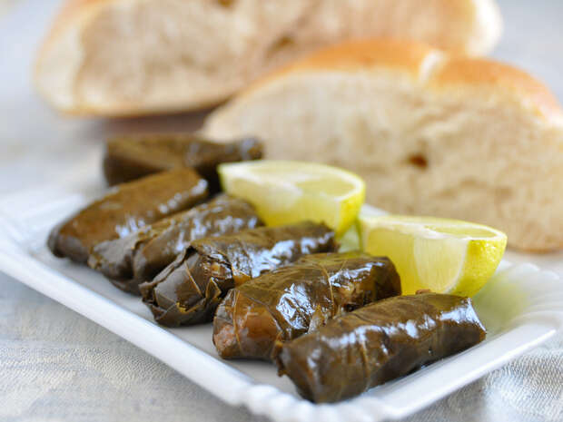 Dolmas rest on a rectangular white plate alongside lemon wedges, set on a tablecloth. In the background, sliced bread adds to the appetizing scene.