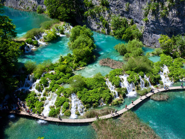 A series of cascading waterfalls flows into turquoise pools, surrounded by lush greenery. A wooden boardwalk with people walking crosses the water, set against rock cliffs in a natural landscape.