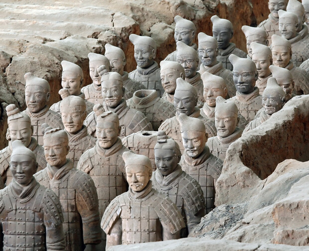 Terracotta soldiers stand silently, lined up in rows. These clay figures wear armor and unique headgear, set within an archaeological excavation site of earthy, dusty trenches and surrounding soil.