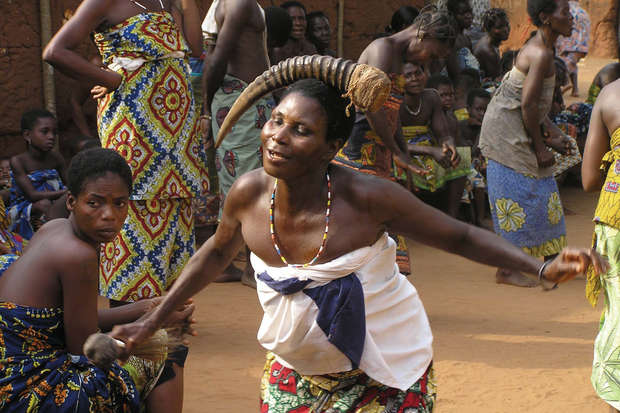 A person dances energetically, adorned with a headdress and traditional clothing. People in colorful attire sit and stand around, observing in a communal, open-air setting.