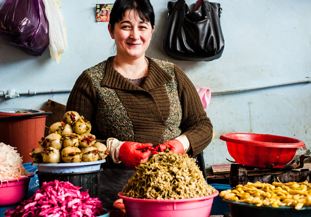 A smiling woman wearing a brown sweater prepares food at a market stall. Various bowls with pickled vegetables and bright colors surround her, set against a light blue wall with hanging bags.