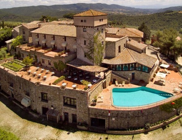 A historic stone building with a central tower overlooks a turquoise swimming pool, surrounded by terracotta patios and hills in the background, creating a tranquil countryside setting.