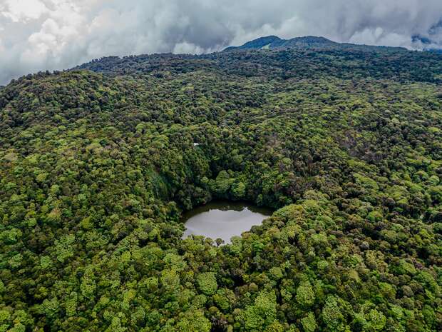 A circular pond rests amid dense, lush green forested hills under a cloudy sky, creating a sense of secluded tranquility in a vibrant natural landscape.