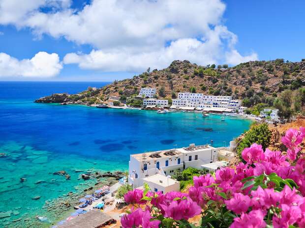Pink flowers bloom in the foreground overlooking azure waters and a rocky coastline lined with white buildings under a bright blue sky with clouds, creating a picturesque coastal scene.