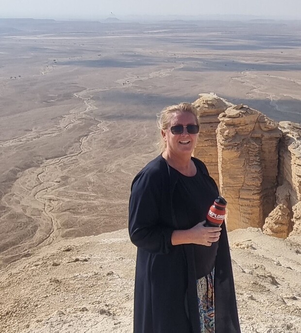 A person in sunglasses smiles while holding a red bottle labeled "EXPLORE" on a rocky cliff, overlooking a vast, arid desert landscape with distant hills.