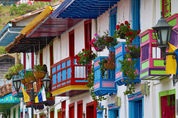 Colorful balconies adorned with blooming flowers overlook a narrow street, with vibrant facades featuring shades of blue, red, and green. Lanterns and hanging plants add charm to the scene.