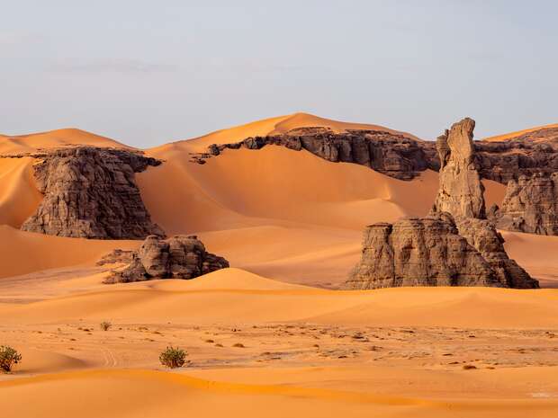 Rugged rock formations stand amidst vast, undulating sand dunes in a desert landscape, displaying shades of orange and brown under soft, natural light, with sparse vegetation dotting the sandy foreground.