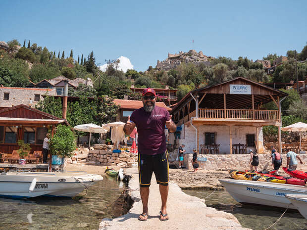 A person stands smiling and giving thumbs up on a stone pathway by a shoreline. Nearby boats rest on clear water. Rustic buildings and lush hills form the background. Sign reads "Kalimche".