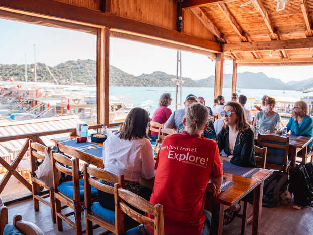 People sit and converse at wooden dining tables inside a restaurant by the sea. Background shows boats, mountains, and blue skies. A red shirt reads, "Don’t just travel. Explore!"