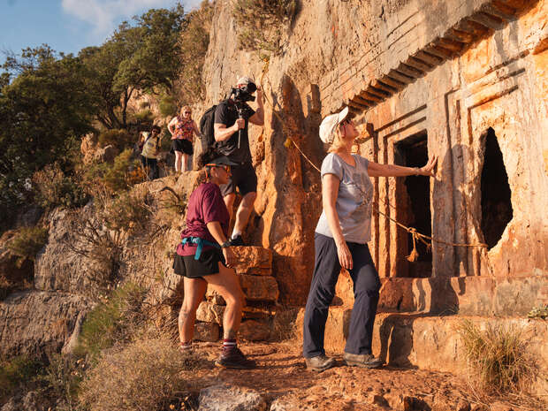 Hikers explore an ancient rock-carved structure on a rocky cliffside, touching the stone facade, with lush trees and a clear sky in the background.