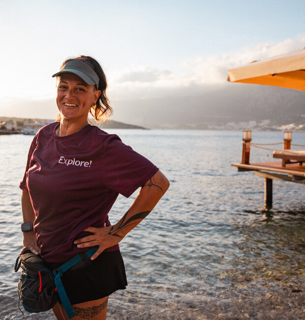 A person wearing a "Explore!" shirt stands smiling with hands on hips by a calm waterfront, under a partly cloudy sky. They wear a visor and a smartwatch, with distant mountains visible.