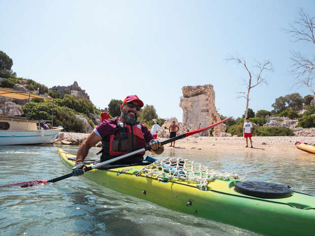 A man paddles a yellow kayak near a sunny beach. Behind him, people stand on the sandy shore, with a rocky landscape and clear blue sky in the background.