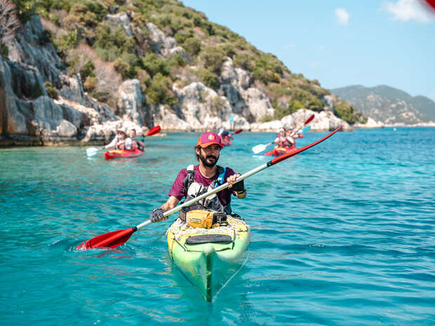 A man paddles a green kayak through clear blue water, followed by others kayaking near rocky, shrub-covered cliffs under a clear sky.