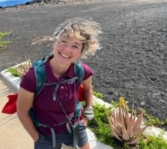 A person with a backpack smiles while standing on a paved path beside a sandy, plant-lined area. The background includes sparse greenery and a distant shoreline.