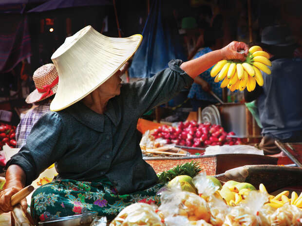 A vendor wearing a wide-brimmed hat holds a bunch of bananas, surrounded by various fruits at a bustling outdoor market with colorful produce and other vendors in the background.