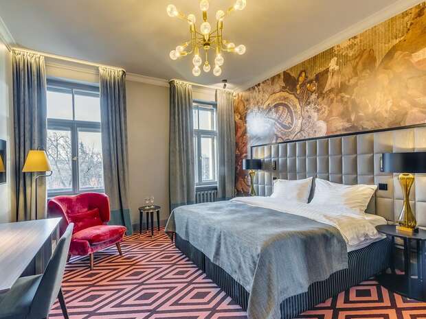 A stylish bedroom features a large, neatly made bed with a quilted headboard beneath a decorative mural. Adjacent, a red velvet chair complements the geometric-patterned carpet, soft lighting, and large windows.