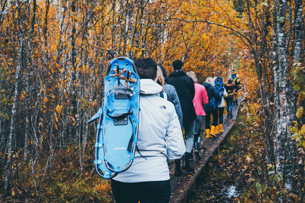 People walk along a narrow wooden path through a forest during autumn. A person in the foreground carries blue snowshoes on their back, surrounded by trees with orange and yellow leaves.