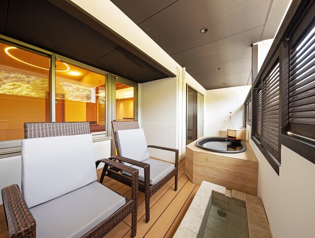 Two wicker chairs face a wooden jacuzzi on a modern balcony with partial walls and windows. The area features a mix of warm lighting and outdoor elements, creating a cozy atmosphere.