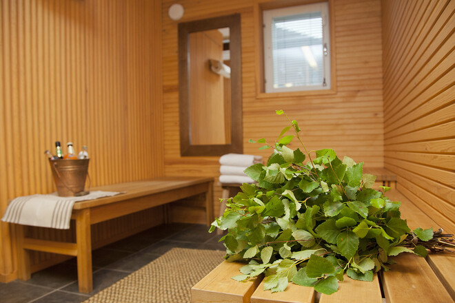 A bundle of green birch leaves rests on a wooden bench in a sauna room, with a bucket and towels on another bench and a small window in the background.