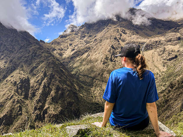 A person in a blue shirt and black cap sits on a grassy edge, facing majestic, rugged mountains under a partly cloudy sky.