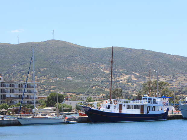 A large blue and white boat docked alongside a smaller sailboat in a tranquil harbor. Surrounding context includes hills in the background and a few buildings near the waterfront.